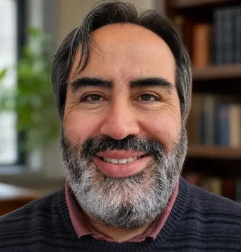 Close-up of a man with dark hair and a gray beard smiling, with a bookshelf in the background.
