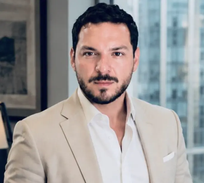 Head-and-shoulders portrait of a man in a beige blazer and white shirt in a modern office setting with a city view behind him.